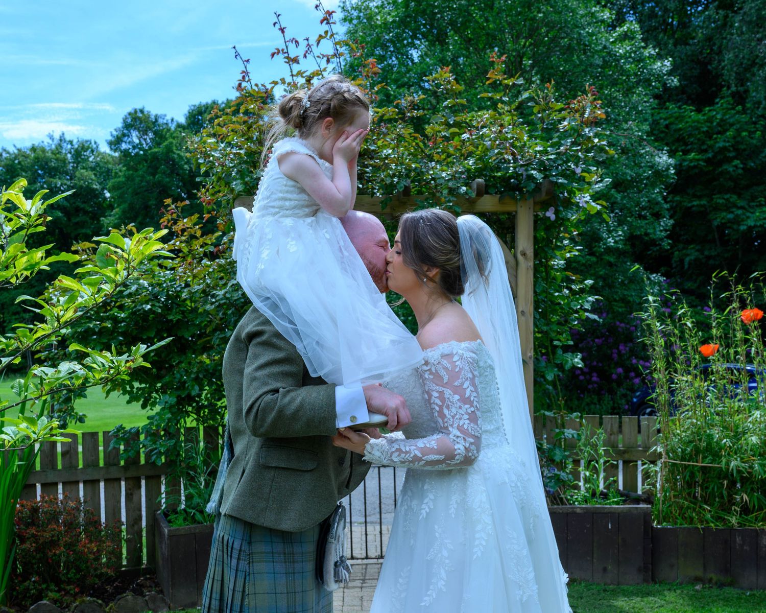 Bride and groom in Scottish tartan kilt share tender moment with flower girl in garden wedding ceremony.