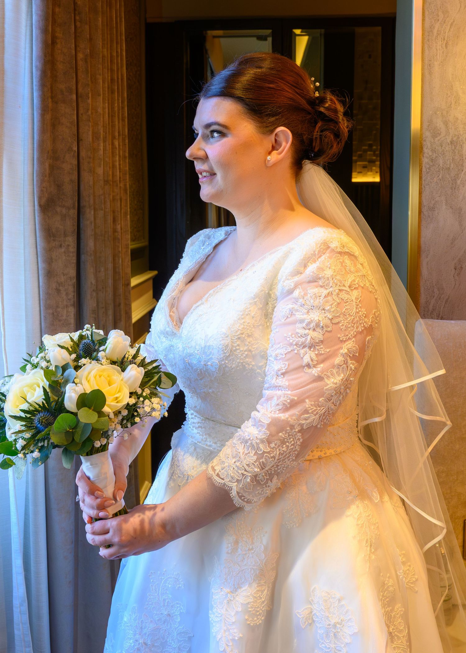 Bride in lace wedding dress with veil holding white rose bouquet, smiling elegantly indoors.