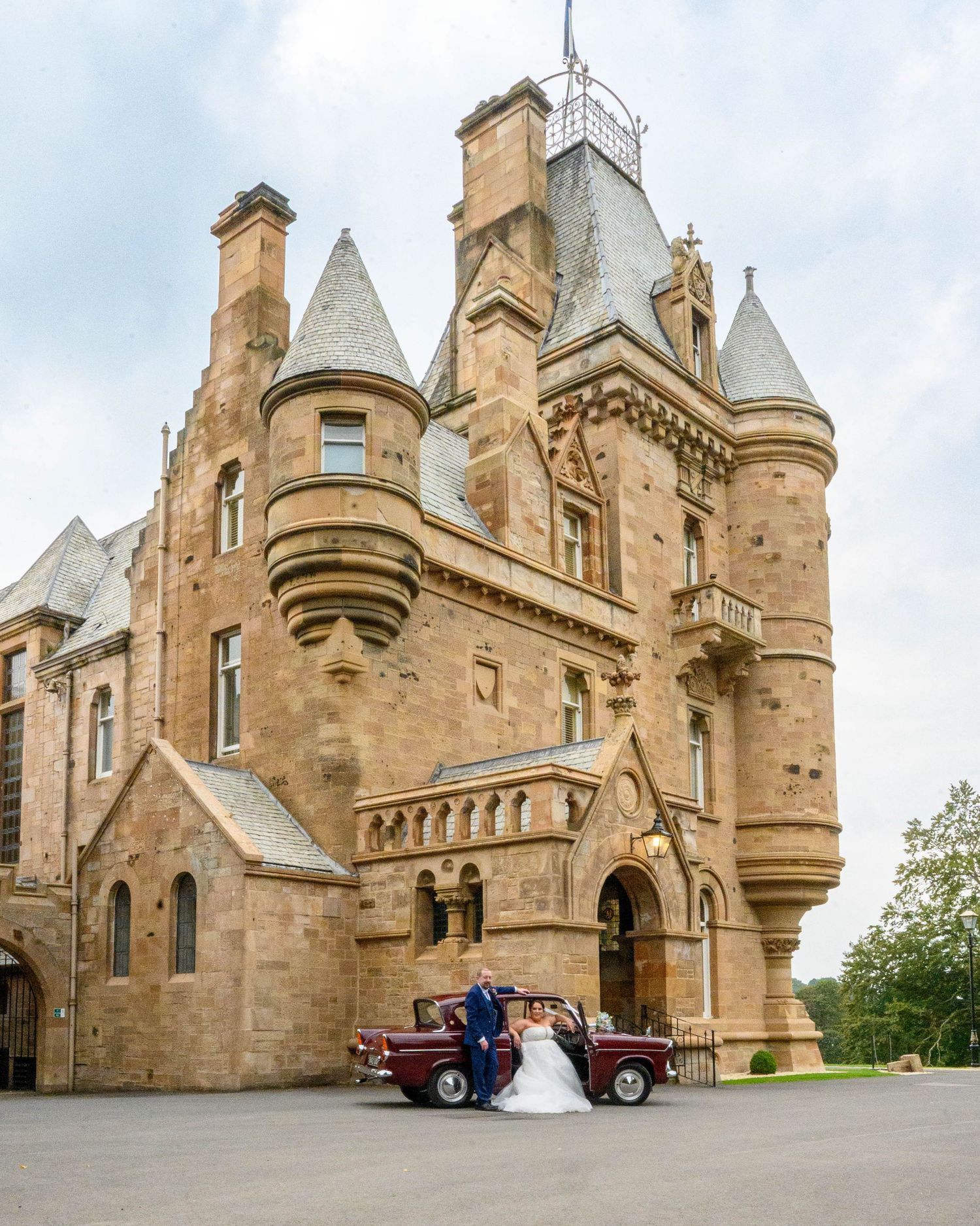 Bride and groom posing with a vintage red car in front of a grand Scottish castle wedding venue.
