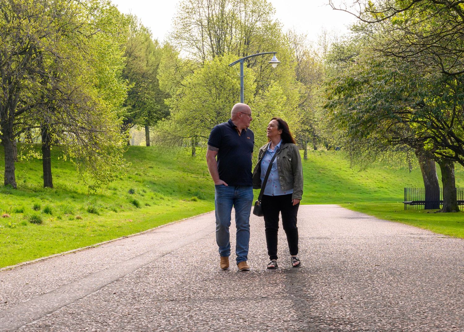 Couple walking together on a tree-lined park path surrounded by lush green grass and trees on a sunny day.