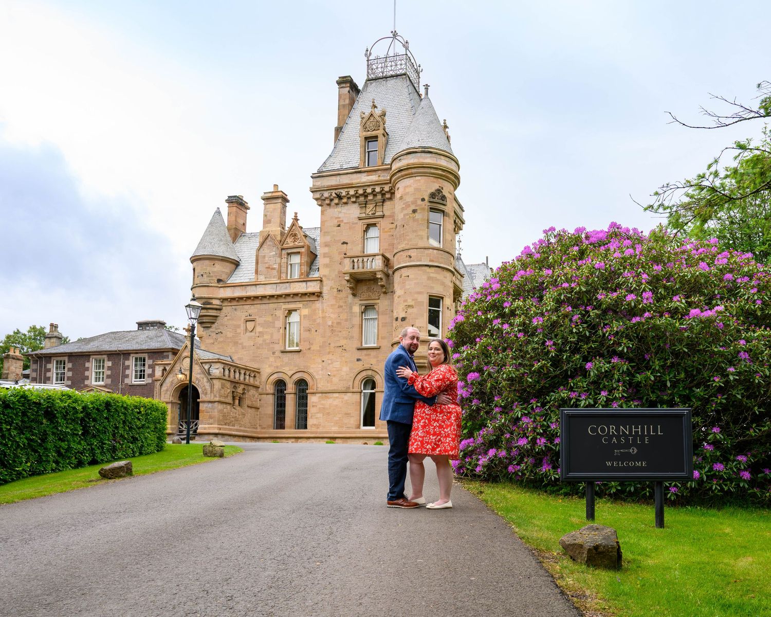 Couple embracing outside Cornhill Castle, a stunning Scottish baronial estate surrounded by lush gardens and blooming rhododendrons.