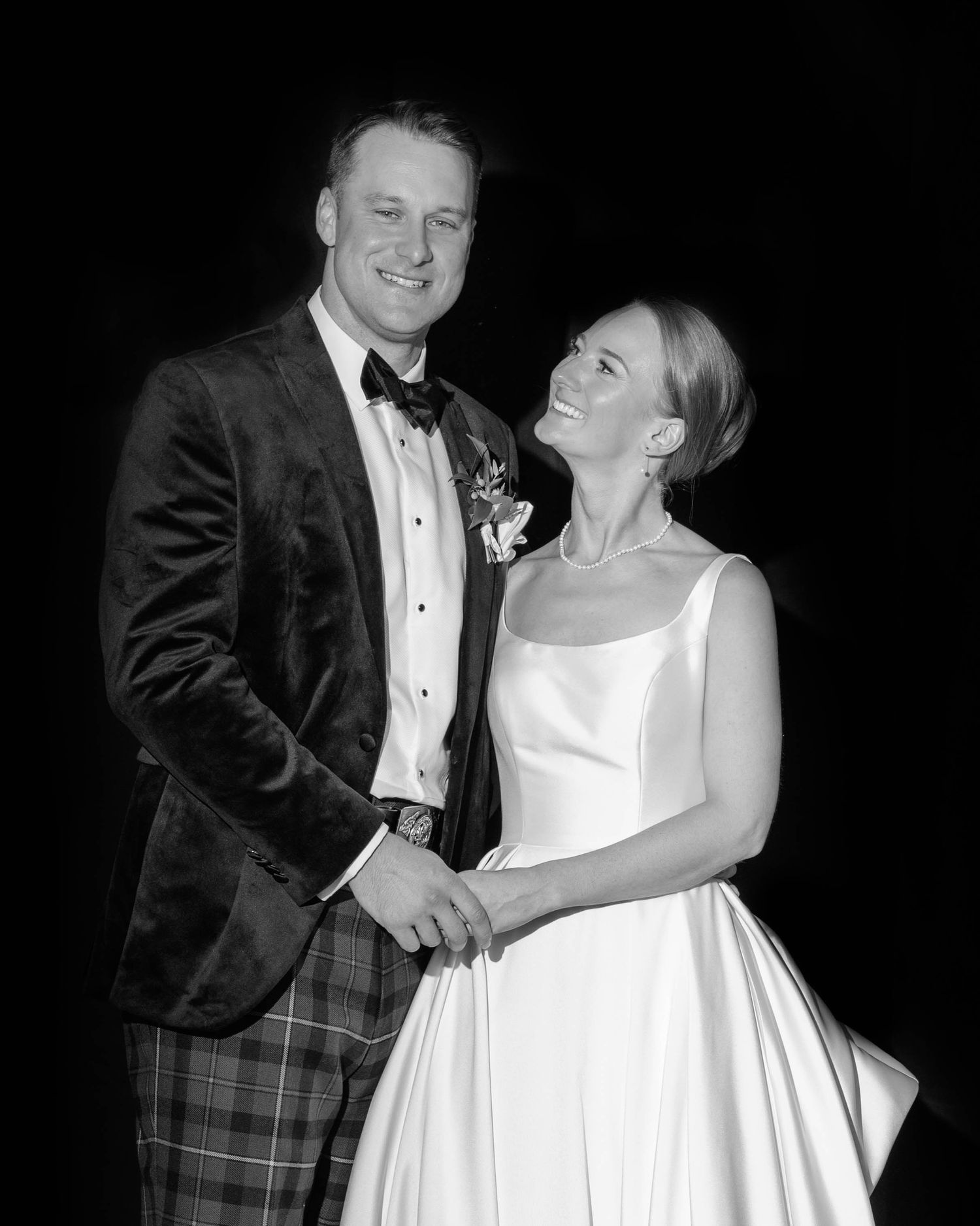 Black and white wedding photo of smiling bride in white gown and groom in plaid tuxedo jacket with bow tie.