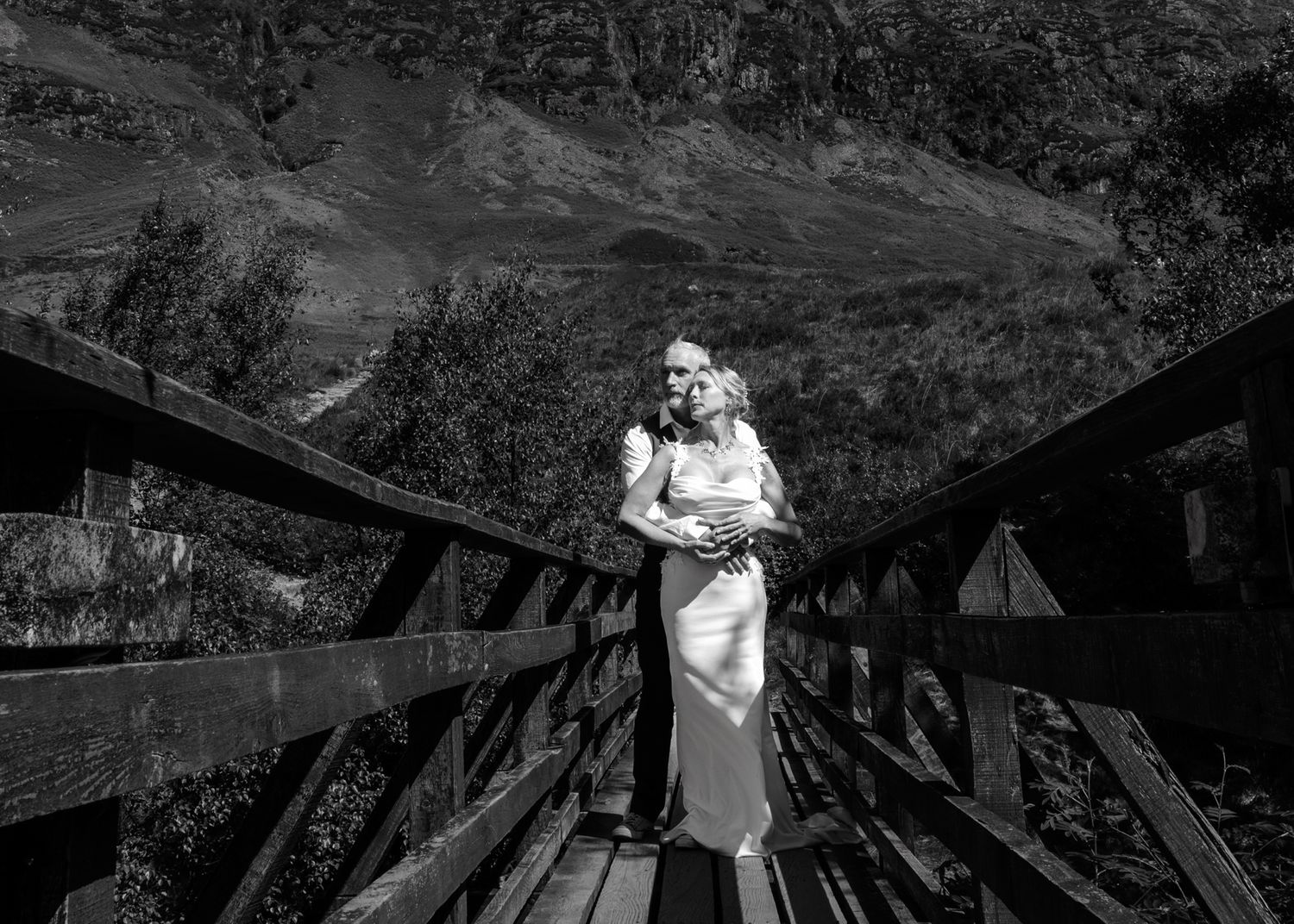 Black and white photo of couple embracing on wooden bridge with mountain backdrop. Glencoe
