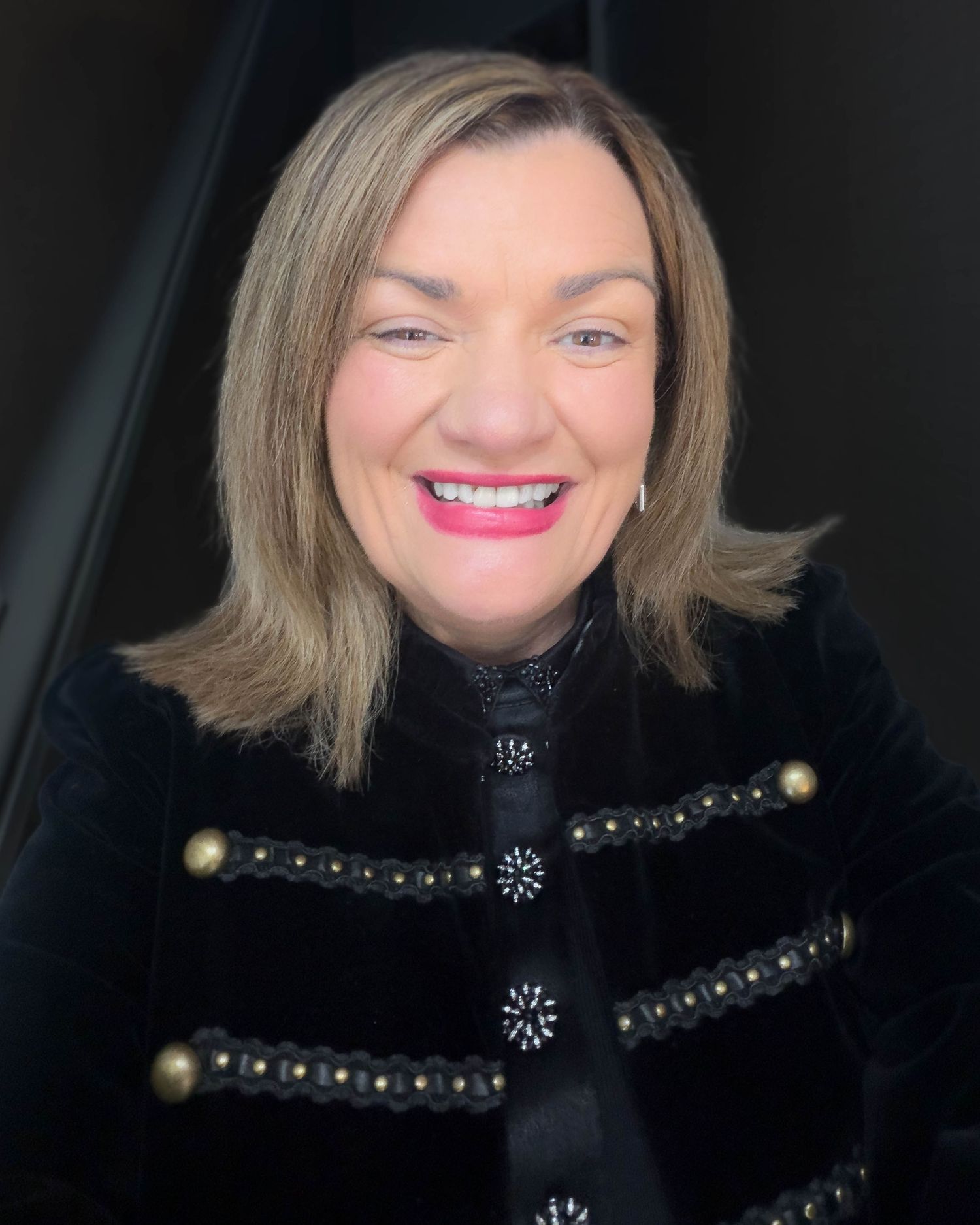 Smiling woman with shoulder-length blonde hair and bright pink lipstick wearing a black velvet military-style jacket with ornate buttons.