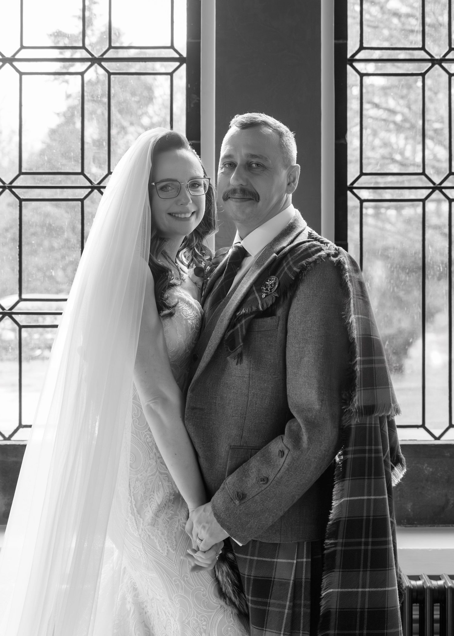 Black and white wedding portrait of a bride in veil and groom in Scottish kilt before a leaded glass window.