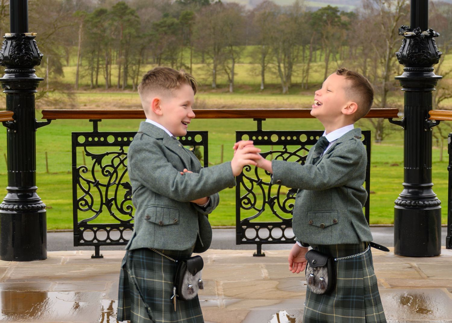 Two people in gray suits playfully dance together on a paved walkway in a park setting with ornate lampposts.