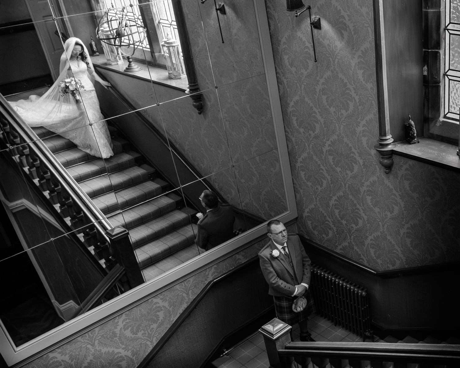 Black and white wedding photo of bride descending grand staircase while groom waits below in elegant venue.