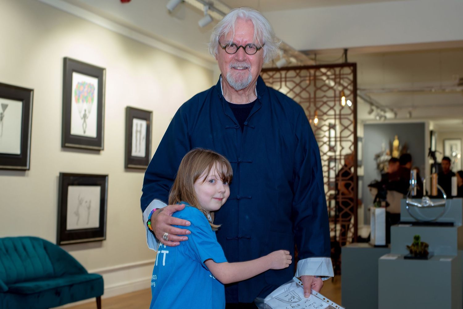 An elderly man with white hair and glasses hugs a young child in a blue shirt inside an art gallery.