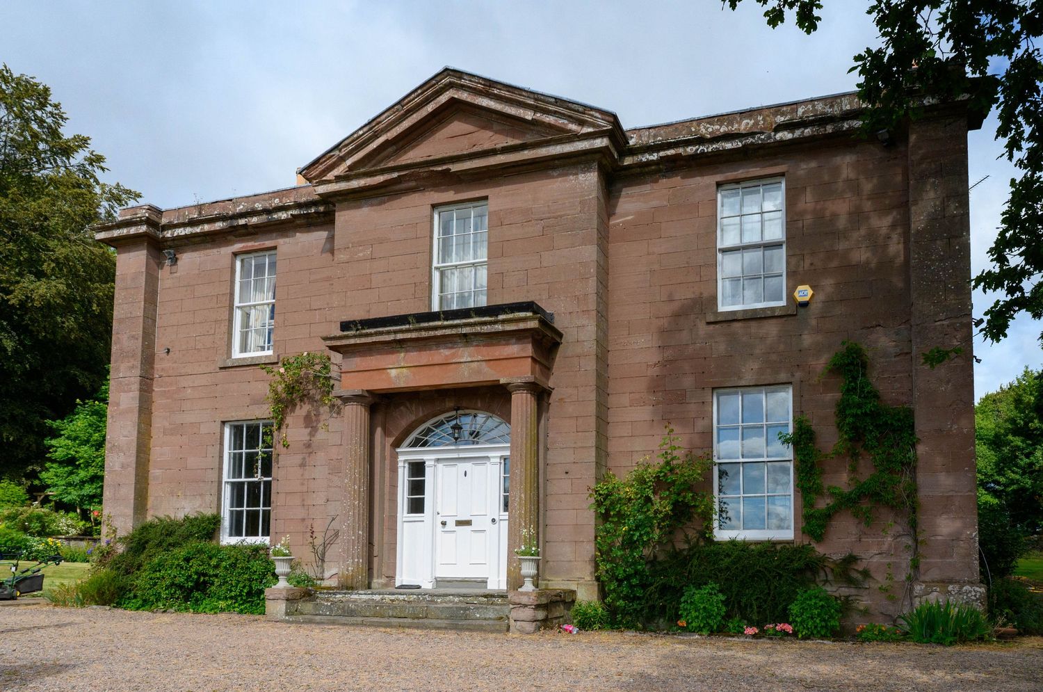 Georgian red sandstone country house with white door, pediment portico, sash windows, and ivy-covered walls surrounded by mature trees.