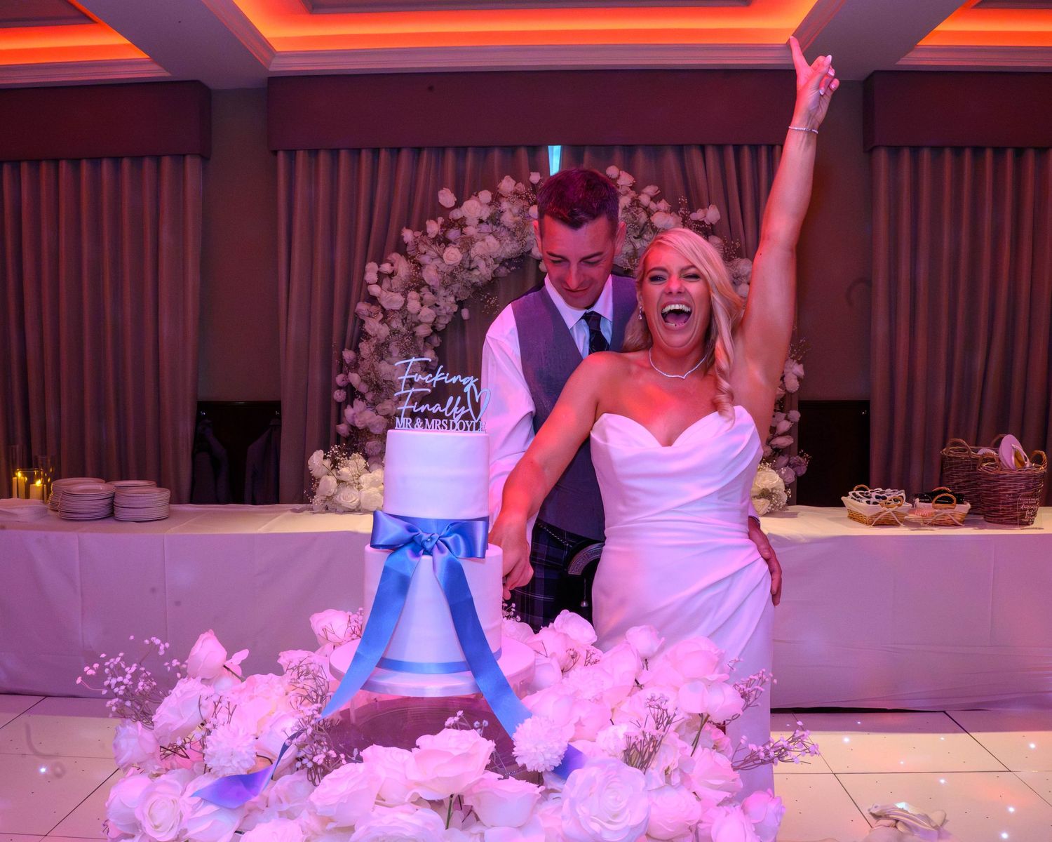 Joyful bride and groom cutting their white and blue wedding cake surrounded by white roses at elegant reception.