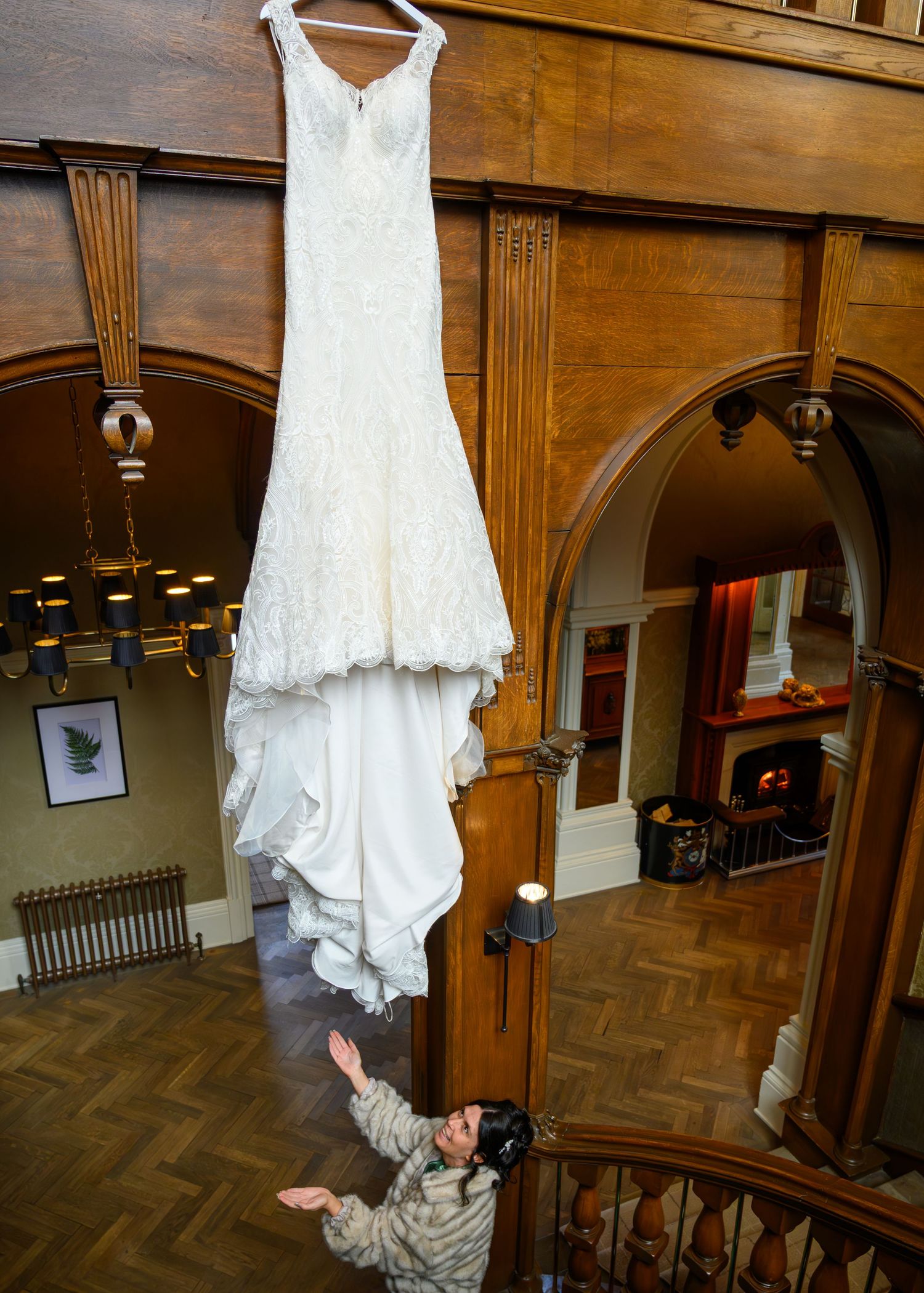 A bride looks up at her elegant white lace wedding dress hanging high in a grand wooden staircase hall.
