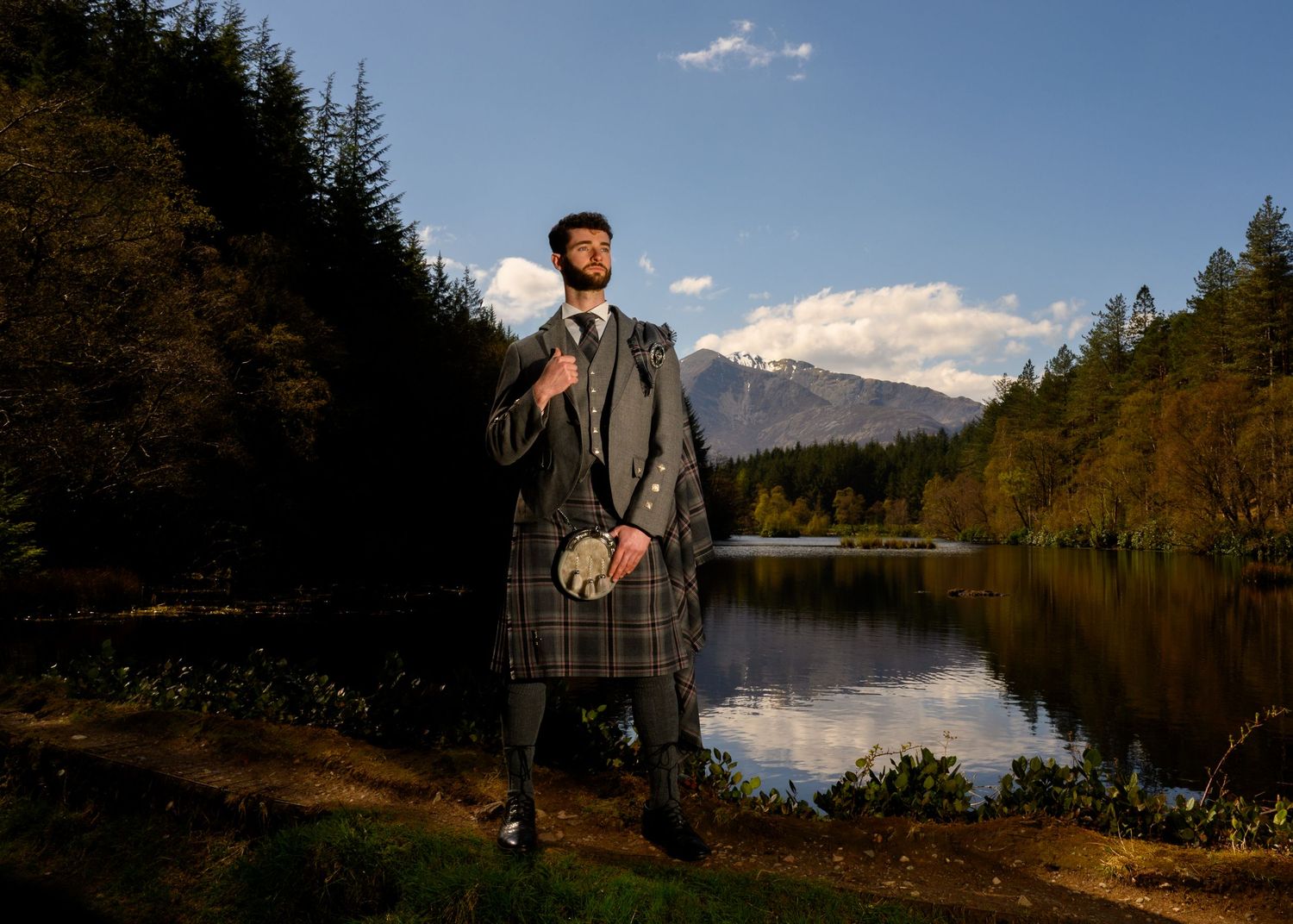 Man in traditional Scottish tartan kilt and jacket stands by a serene loch with mountains and forest backdrop.