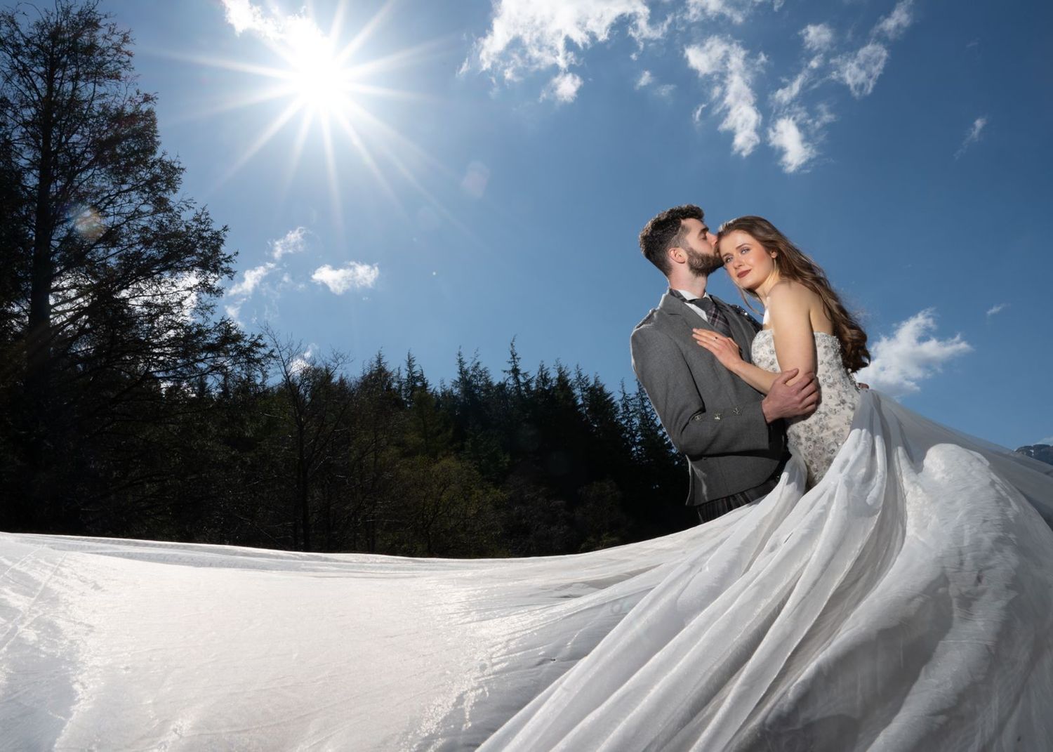 Bride and groom embrace beneath a radiant sunburst with flowing wedding gown against a blue sky.