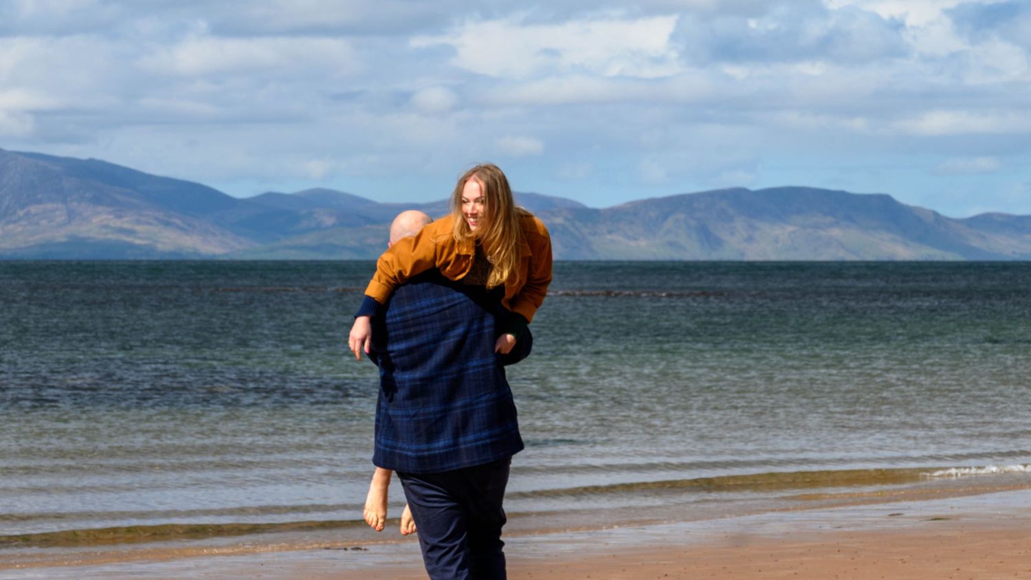 Two red-haired people enjoying a piggyback ride on a scenic beach with mountains in the background.