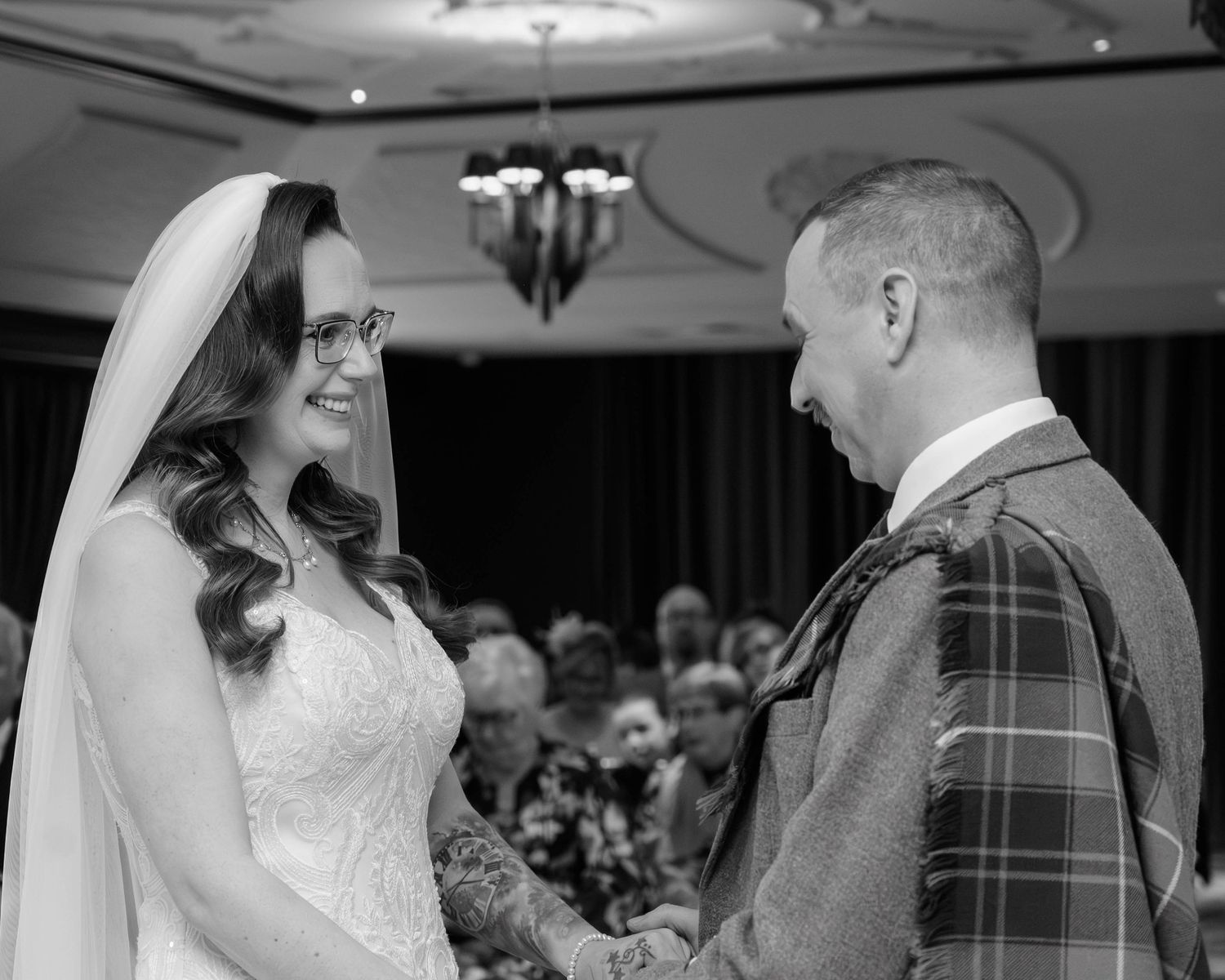 Black and white wedding photo of a smiling bride in veil and groom in tartan kilt exchanging vows at indoor ceremony.