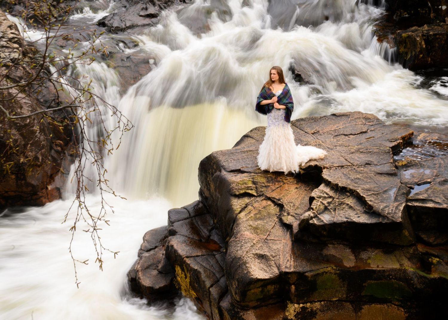 A woman in a white dress stands on rocky terrain before a rushing waterfall in a dramatic outdoor portrait.