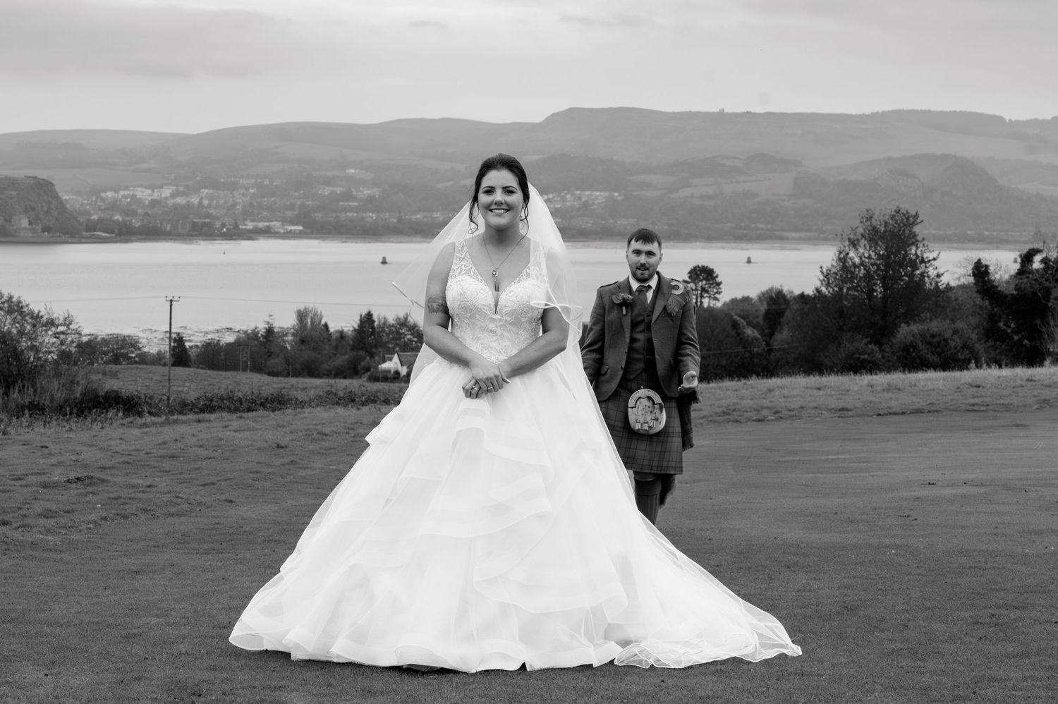 Black and white wedding photo of bride in ball gown and groom in kilt, posing on golf course with scenic loch backdrop.