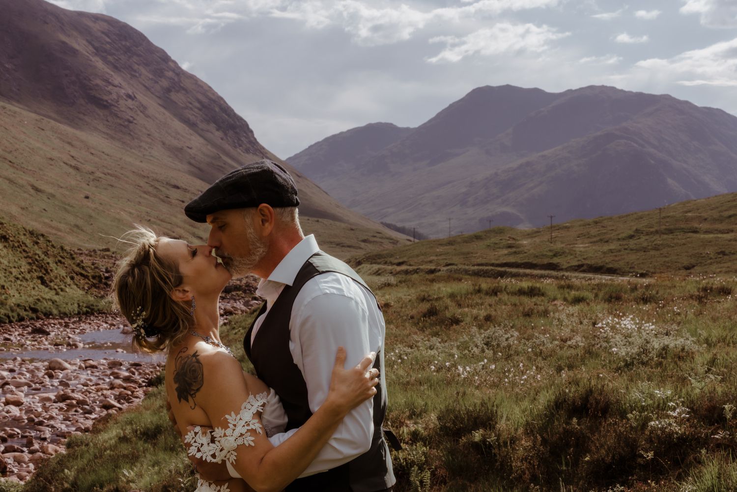 Two people embrace in an intimate moment against the backdrop of scenic Scottish Highlands mountains and valleys. Glencoe
