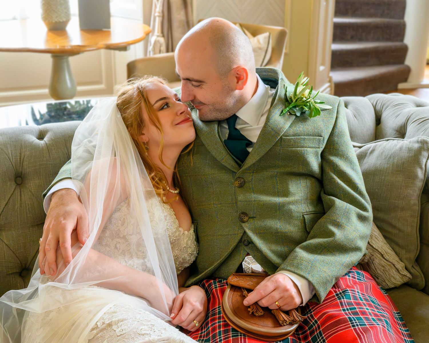 Bride in white veil and groom in tartan kilt share a romantic moment seated on a sofa at a Scottish wedding.