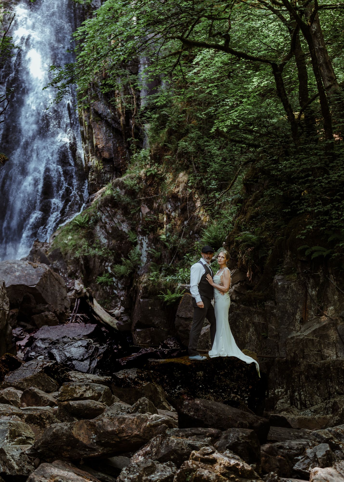 A couple stands together on rocky terrain beside a majestic cascading waterfall surrounded by lush green forest. Glencoe