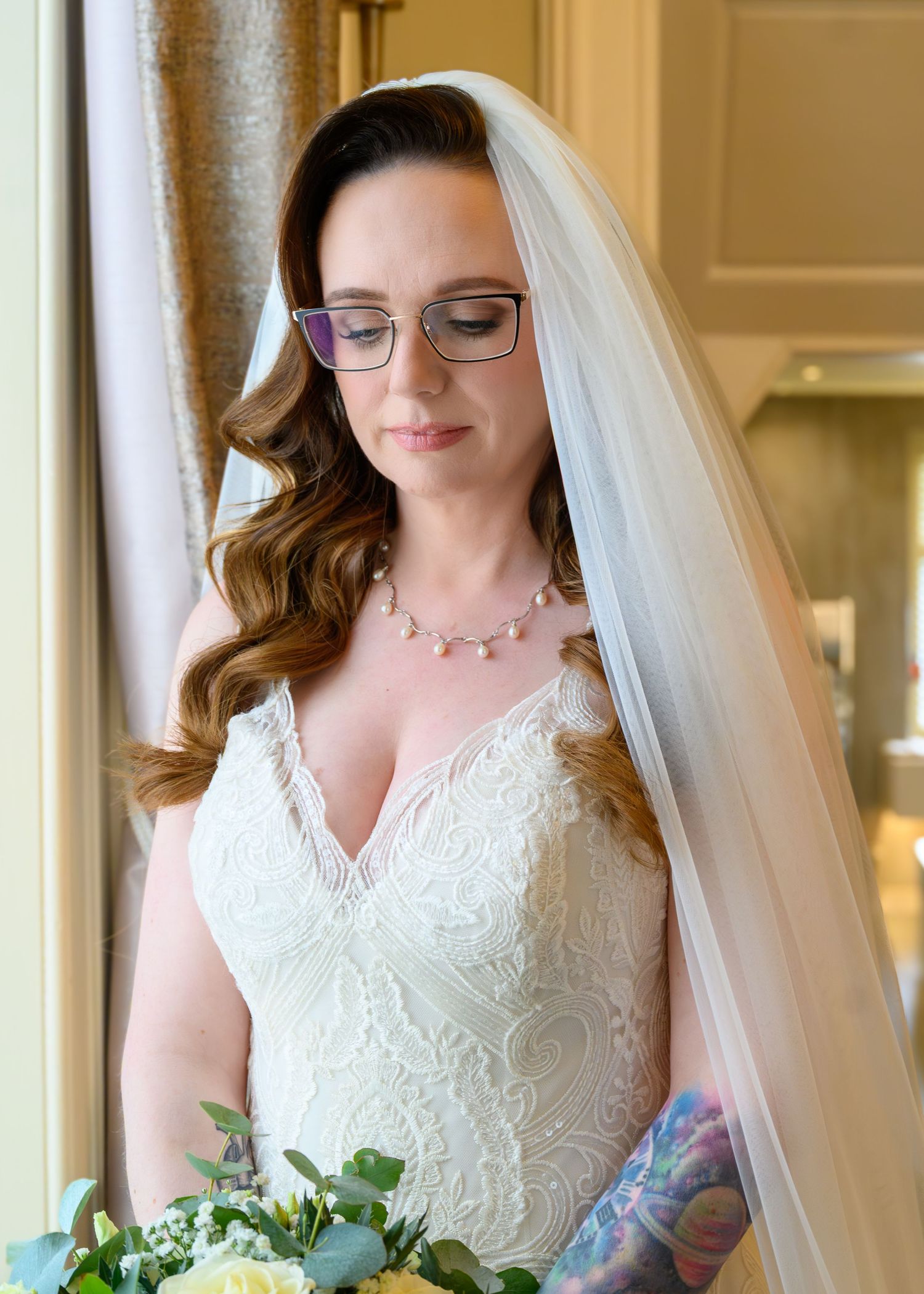 Bride in white lace gown and veil holds bouquet, wearing glasses and pearl necklace, looking down softly.