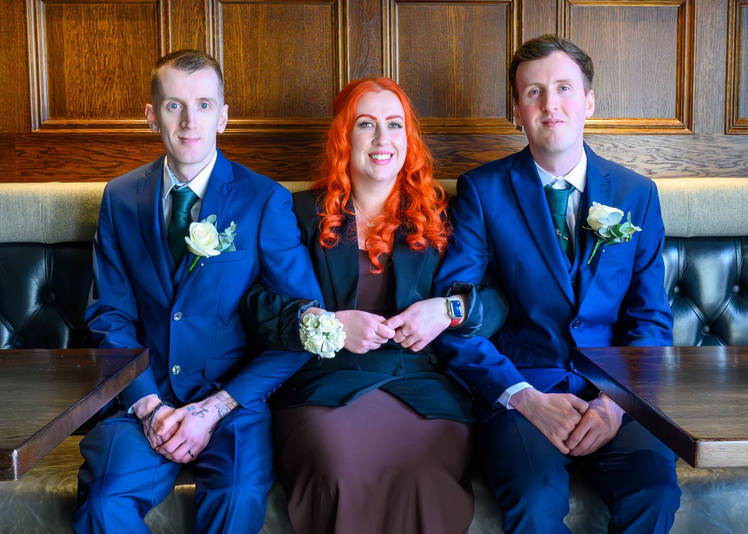 Two men in blue suits and a woman with red hair sit together smiling, wearing floral boutonnieres at a formal event.