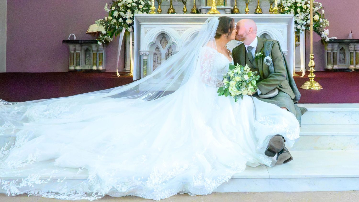 A bride and groom share a romantic kiss at the church altar, her elegant white gown and long veil spread beautifully around them.
