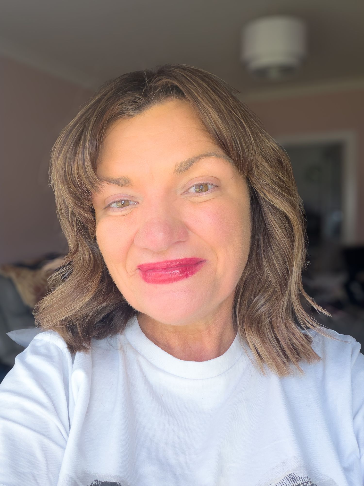 A smiling middle-aged woman with brown shoulder-length hair and red lipstick wearing a white shirt takes a selfie indoors.