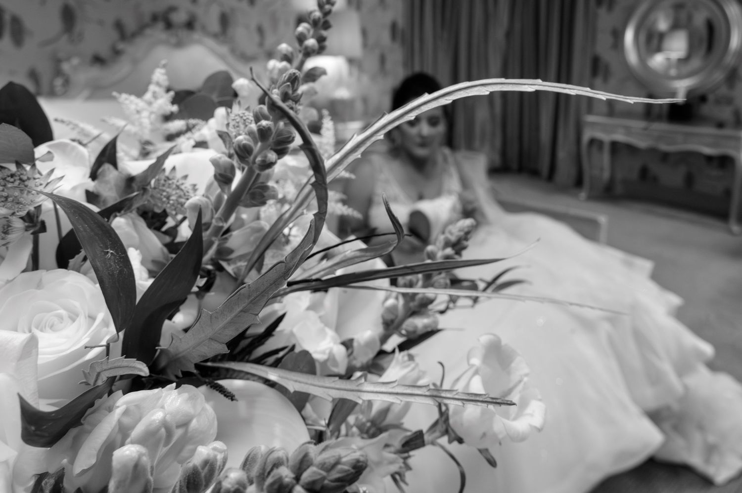Black and white photo of a bride in a wedding dress seated behind an elegant floral arrangement of roses and greenery.