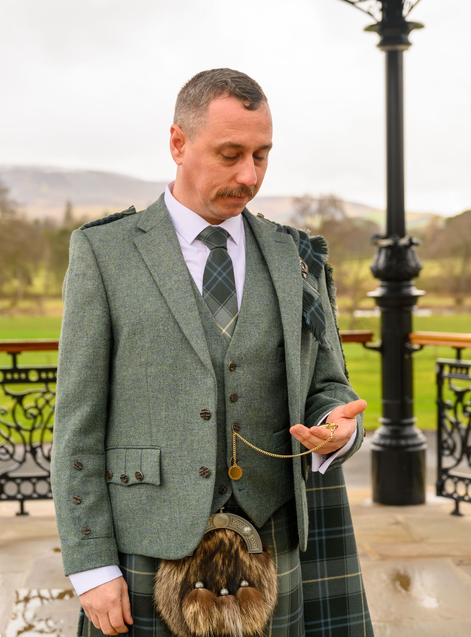 Man in traditional Scottish Highland dress with kilt and tweed jacket holding wedding rings outdoors.