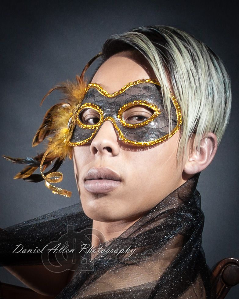 Portrait of person wearing ornate gold and brown masquerade mask with feathers against dark background.