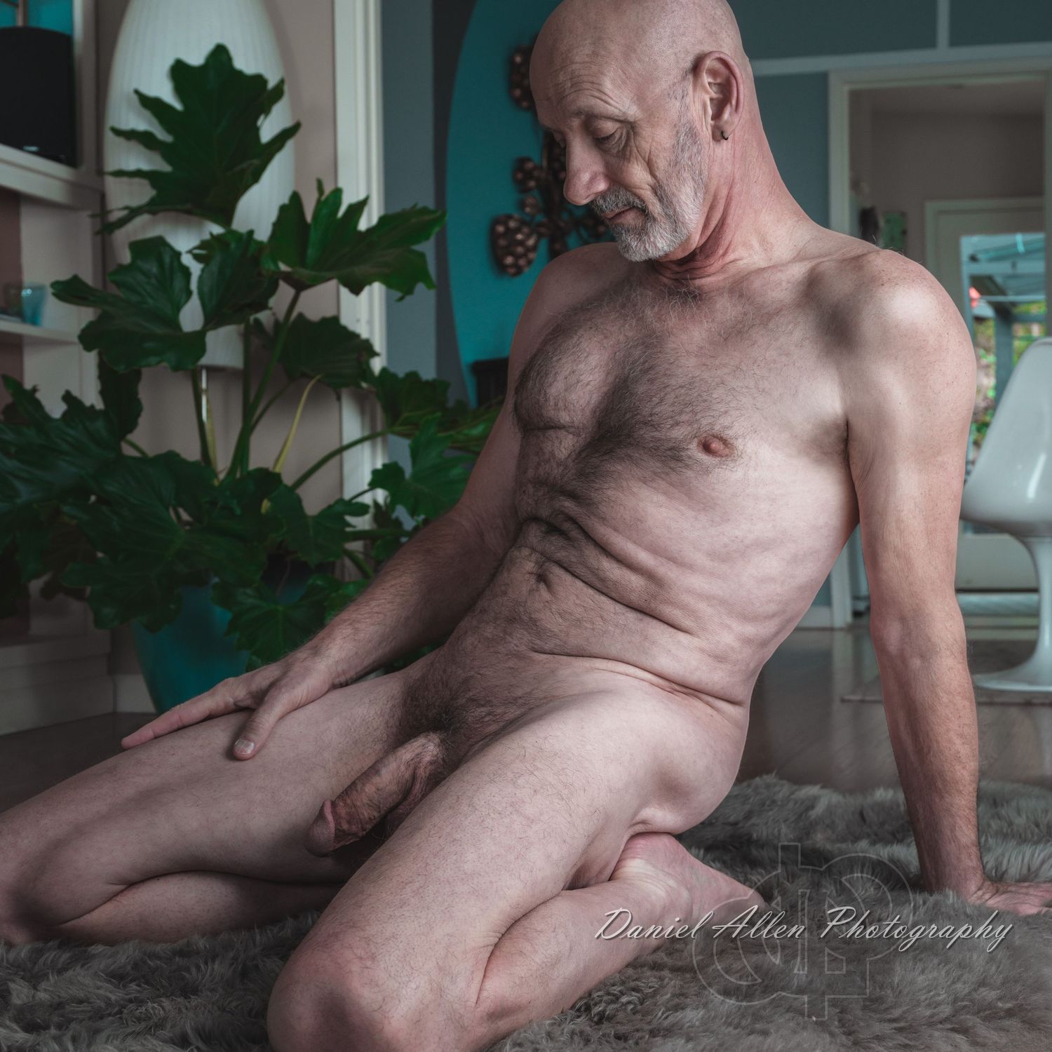 A nude man kneeling on a fur rug on the floor.
