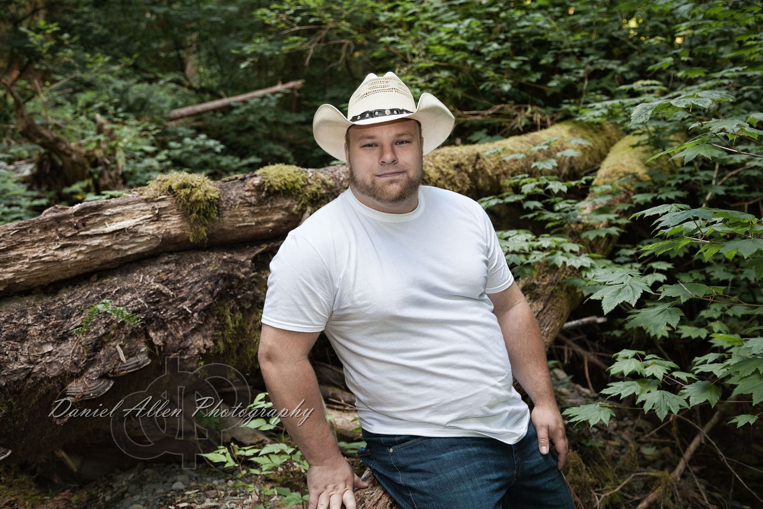 Person in white t-shirt and cowboy hat poses in outdoor wooded setting with fallen logs and green foliage.