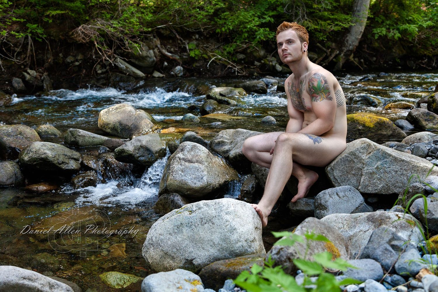 Figure resting on large boulders beside a mountain stream with forest backdrop.