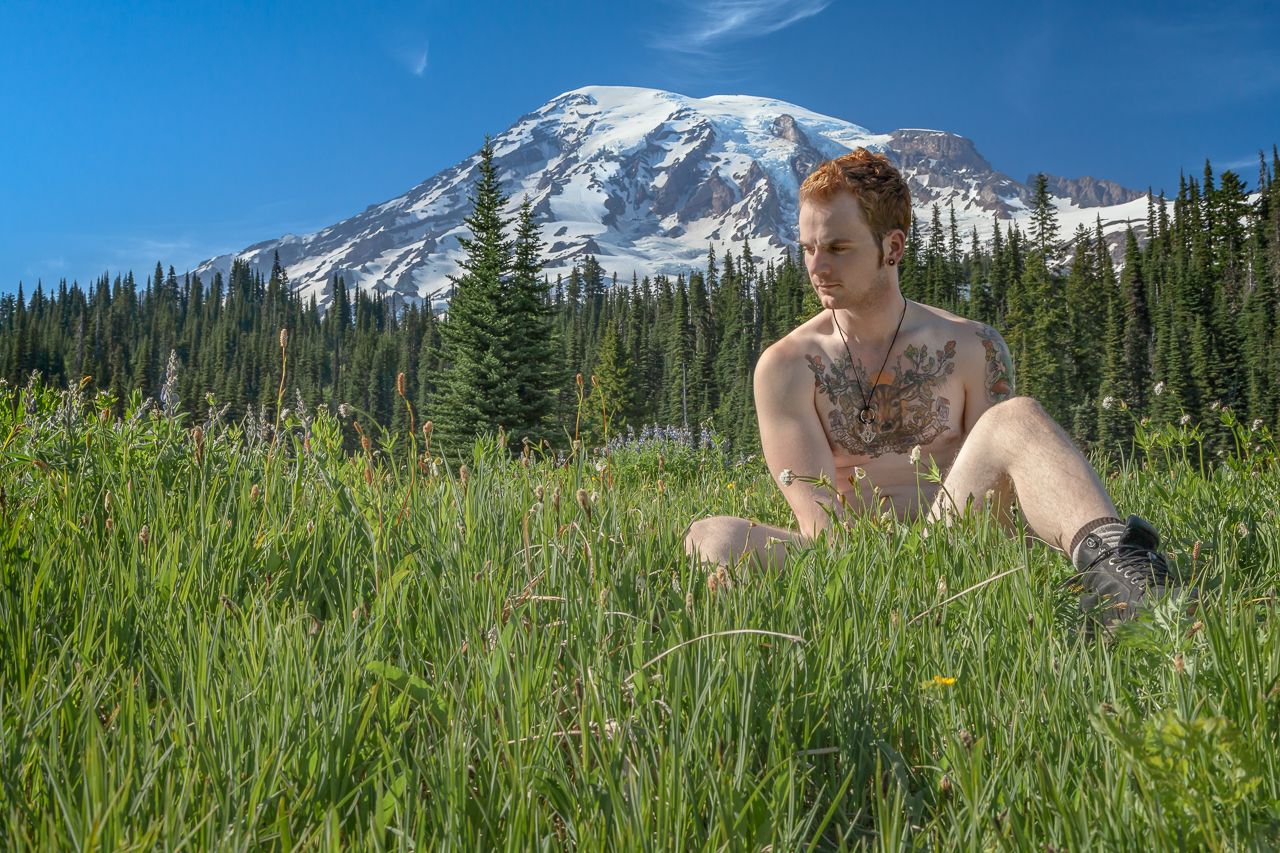 Mount Rainier towers over a nude man sitting in a wildflower meadow on a clear summer day.