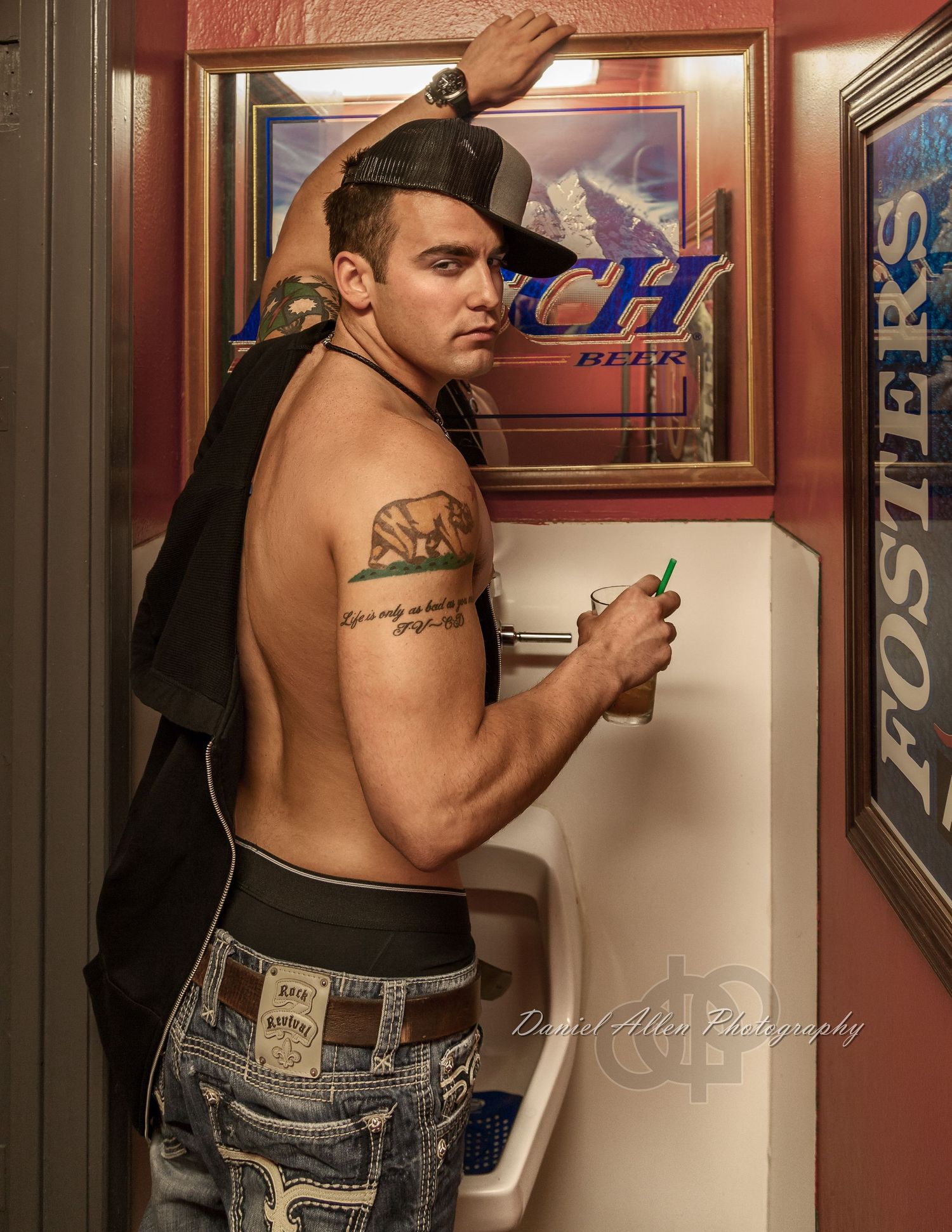 A drunken shirtless man standing at a urinal with a drink in a bar setting.