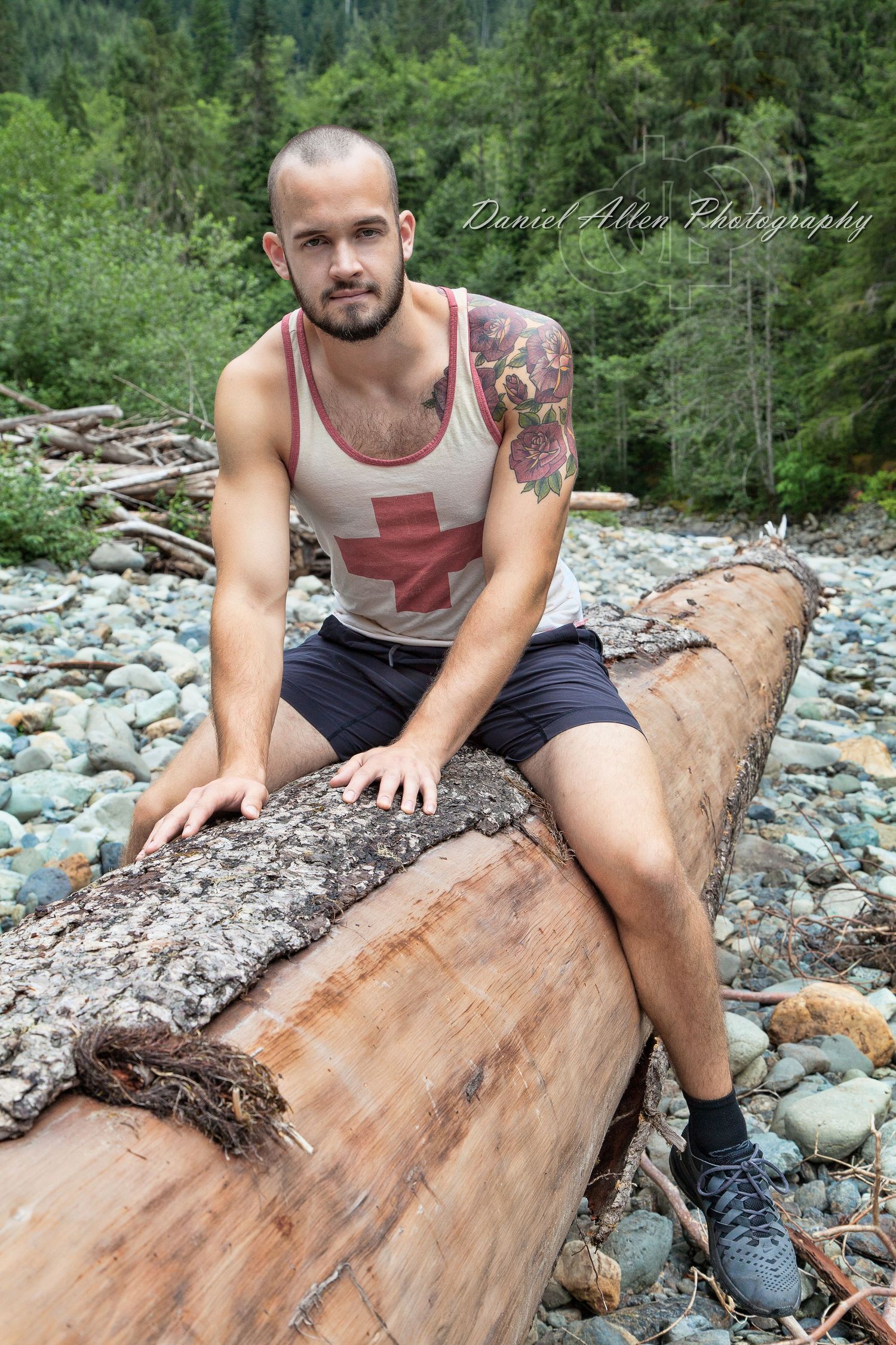 Tattooed person in red cross tank top sits on fallen log by rocky riverbed.