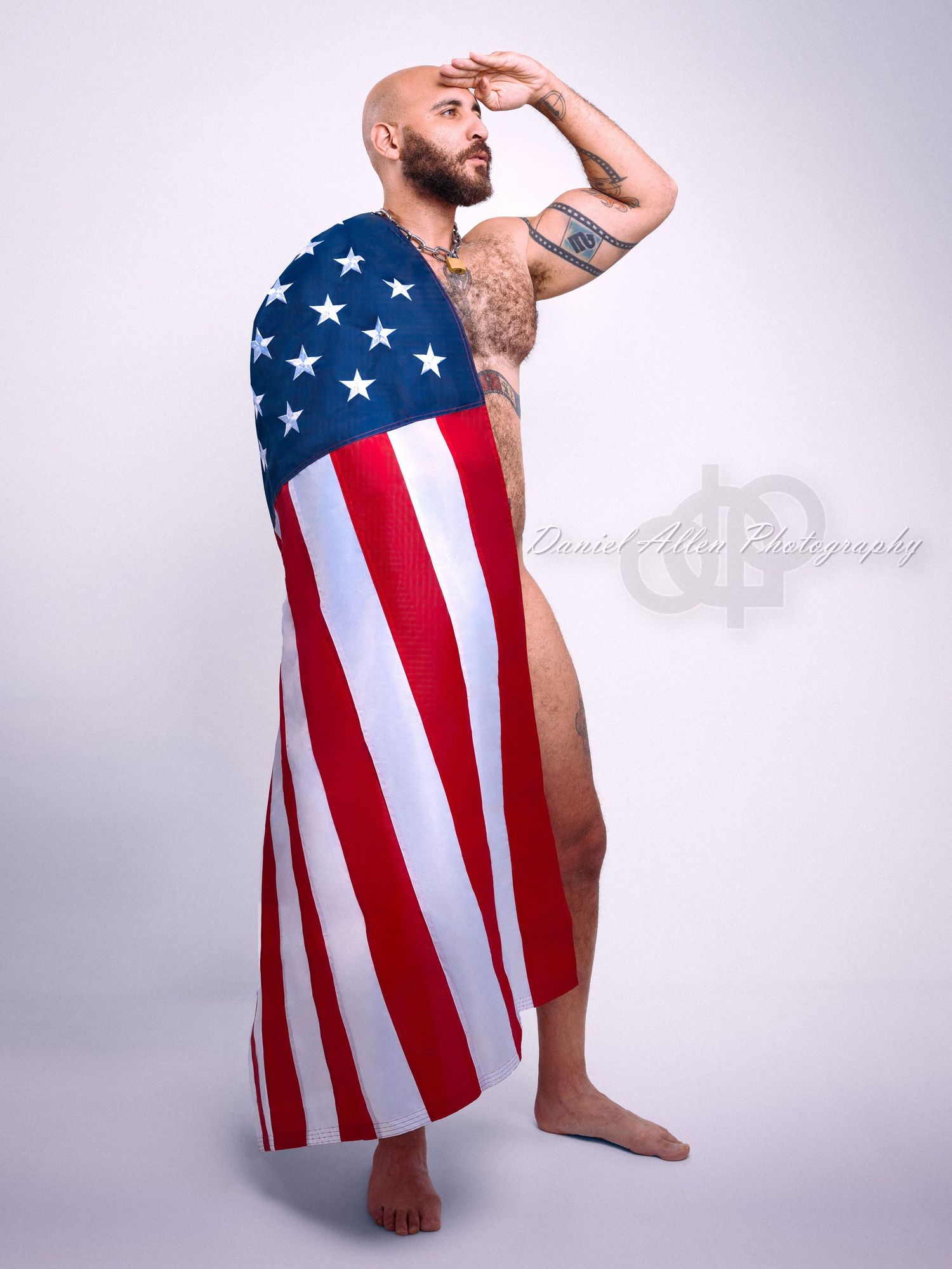 A tattooed figure poses artistically while draped in an American flag against a light gray studio background.