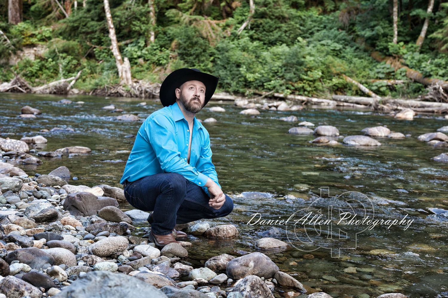 Person in blue shirt sits on rocky riverbank surrounded by flowing water and natural scenery.