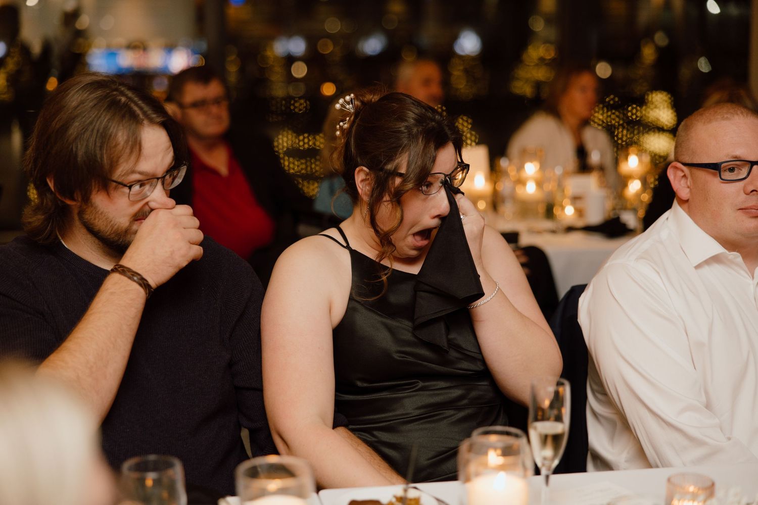 Three guests seated at a dimly lit table during what appears to be an emotional wedding reception moment.