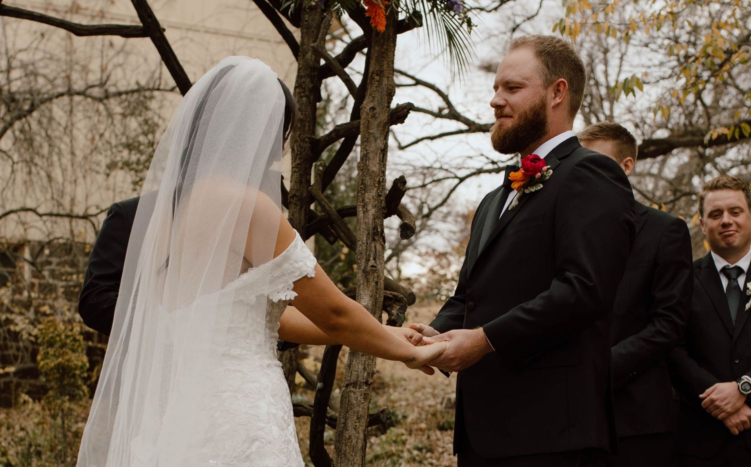 A couple holds hands during an outdoor autumn wedding ceremony in front of bare trees.