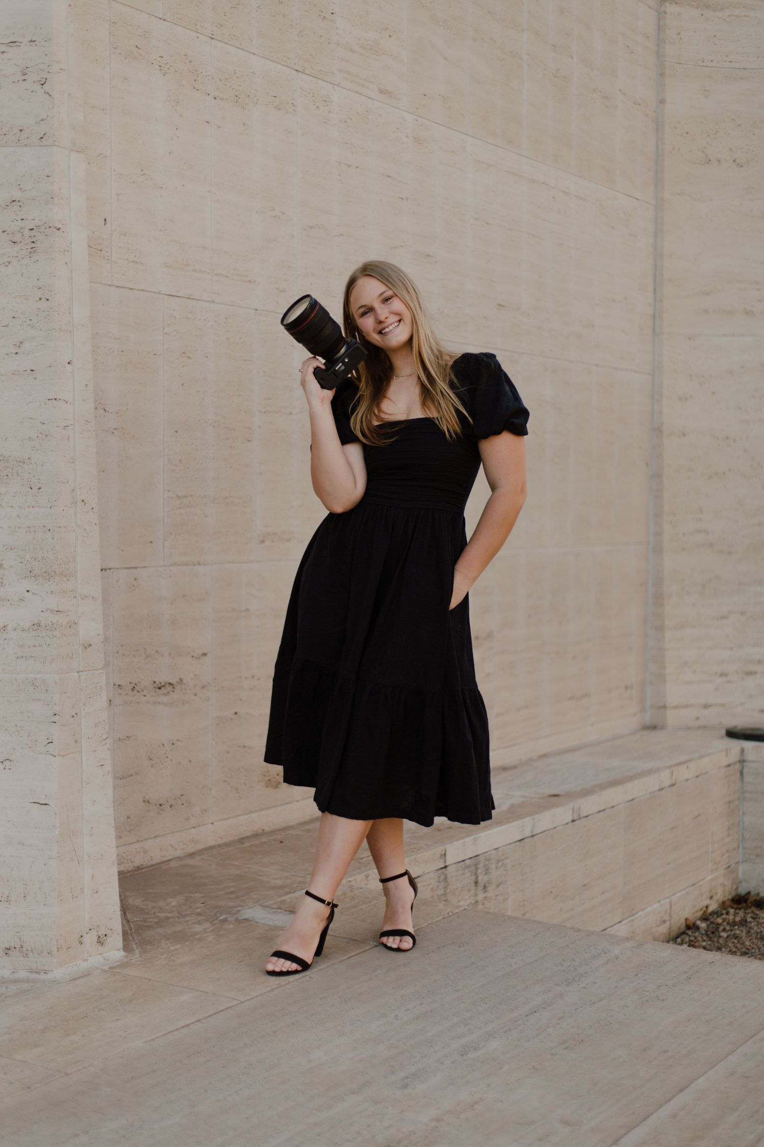 Photographer in black dress poses creatively against beige concrete wall.