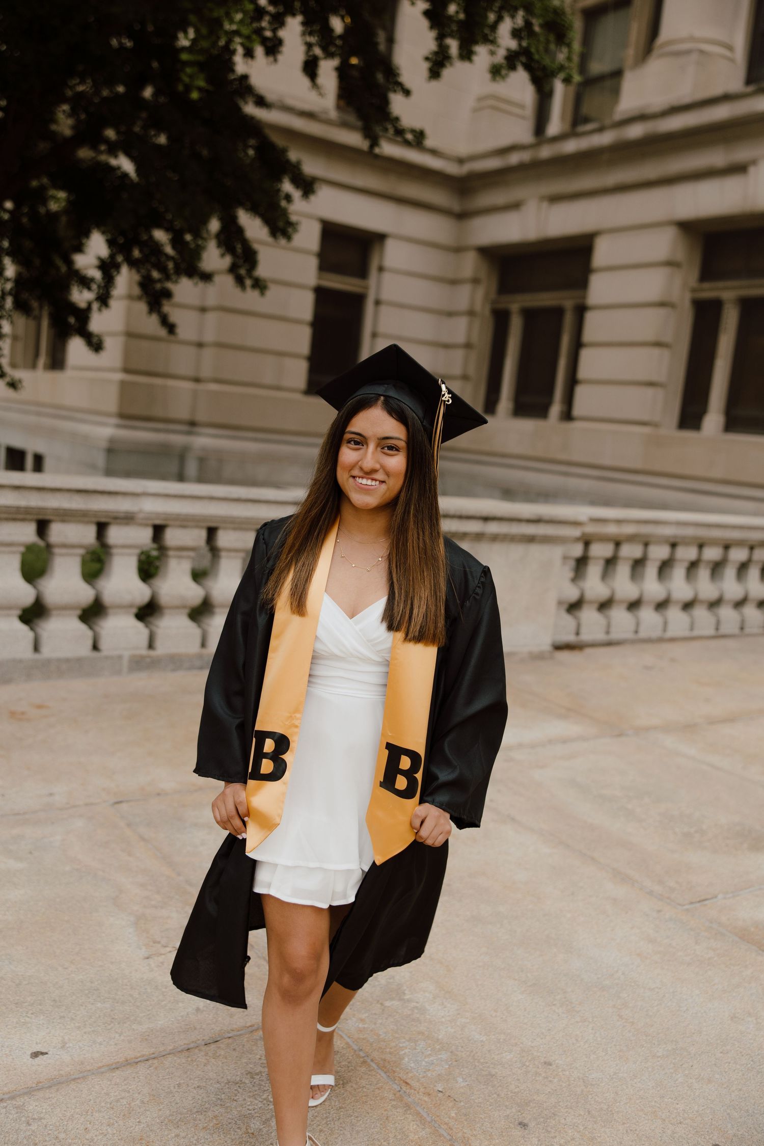 Graduate in black cap and gown poses on campus steps near classical architecture.