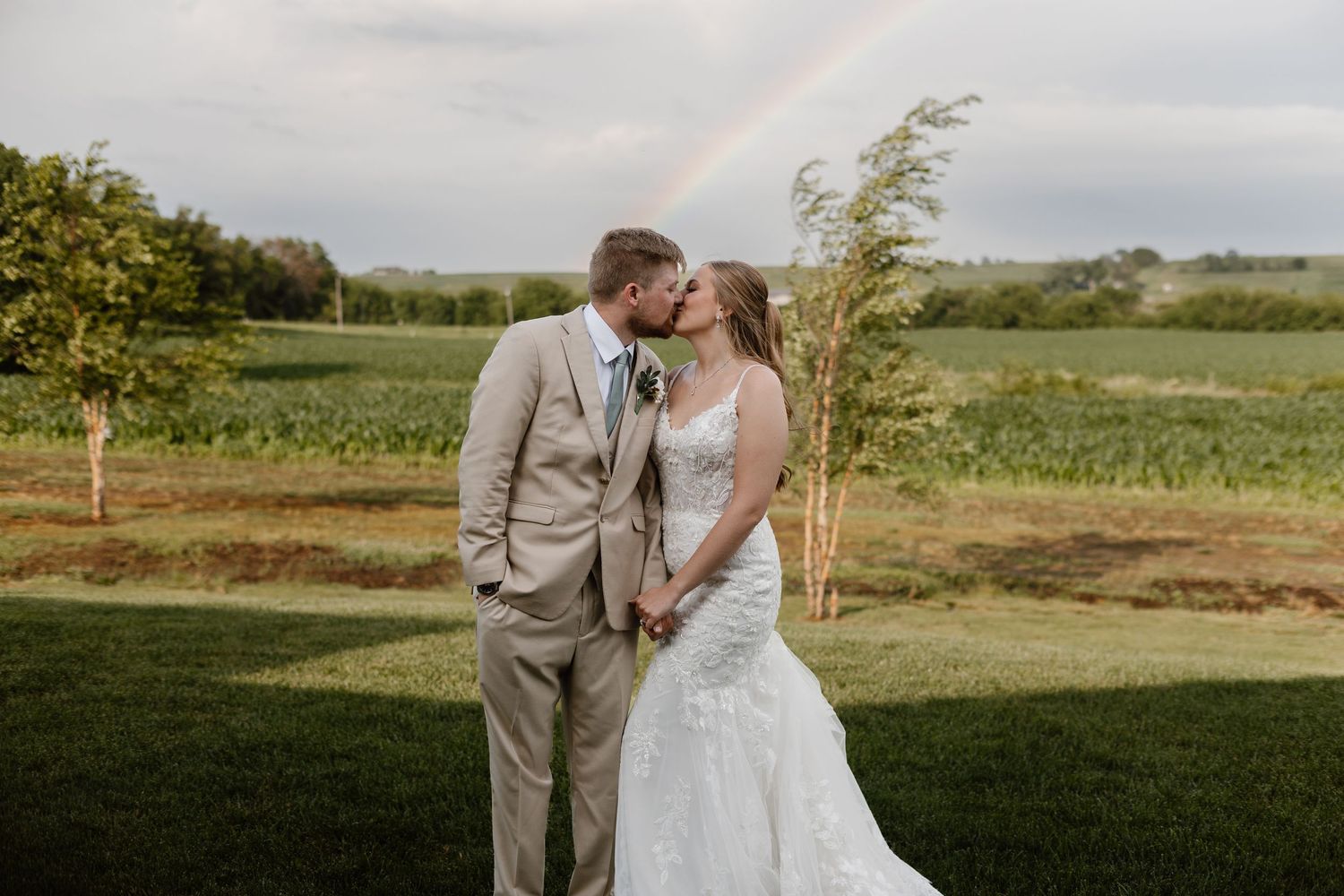 Bride and groom kissing outdoors with a rainbow over green farmland fields in the background.