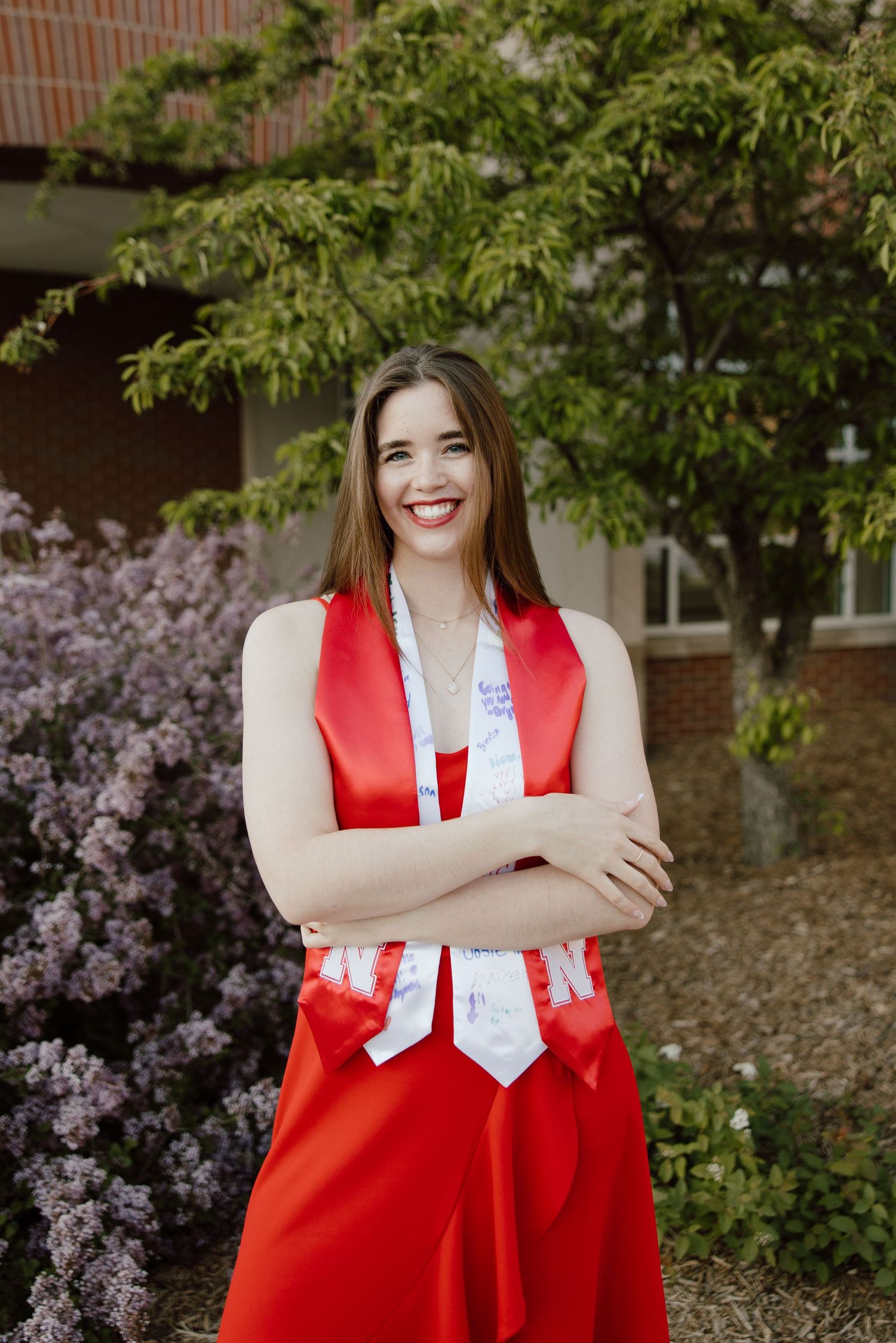 Graduate in red graduation attire smiles during outdoor photoshoot with flowers in background.