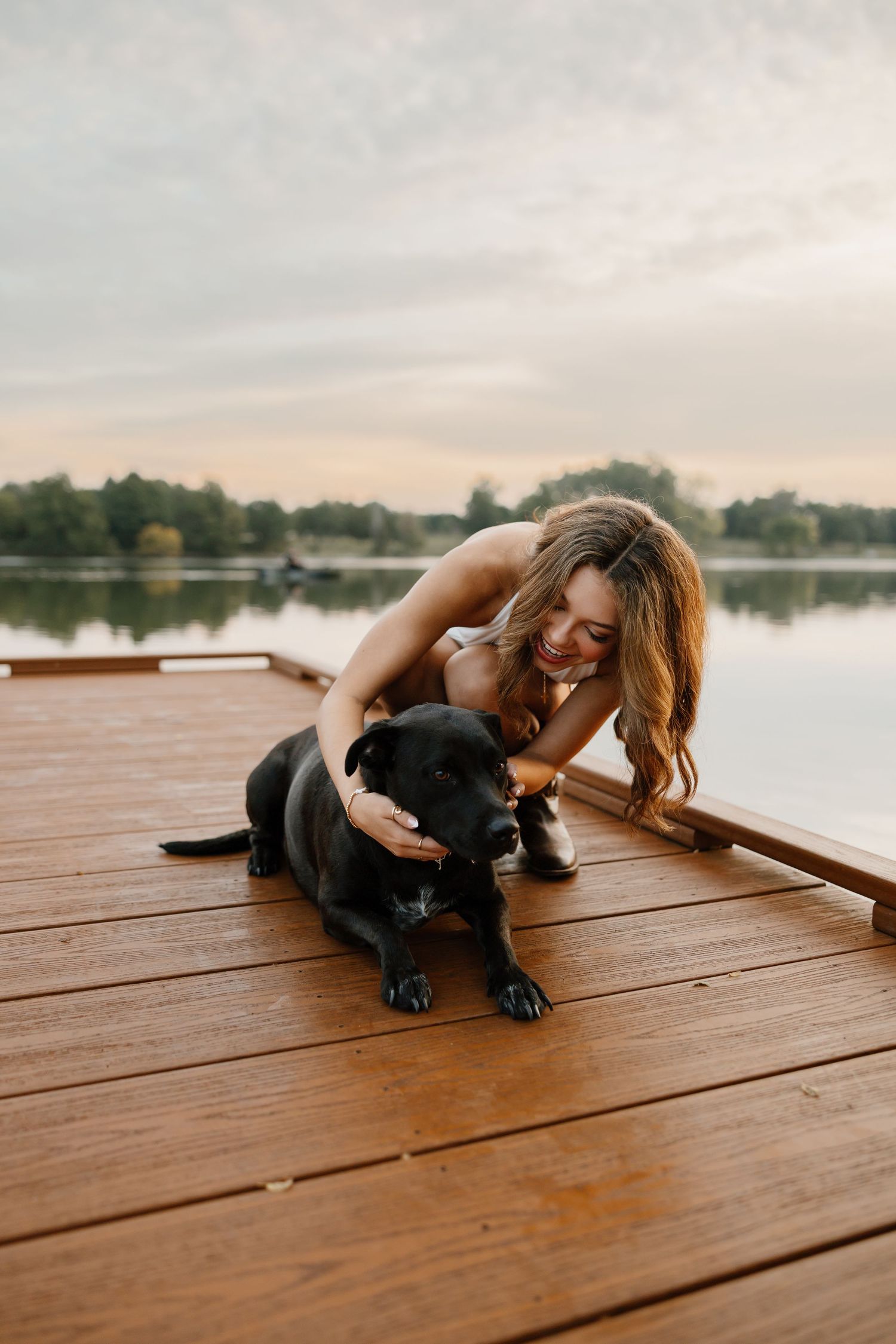 A person lies on a wooden dock playing with a black dog near a serene lake at sunset.