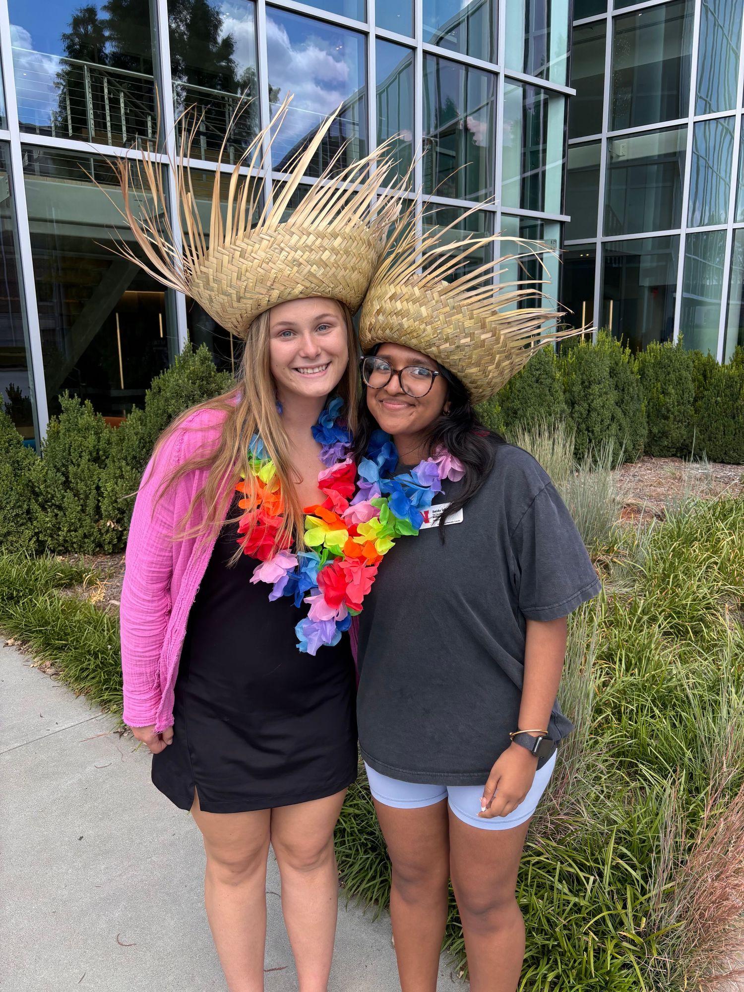 Two people wearing festive straw hats and lei garlands pose outside a modern building.