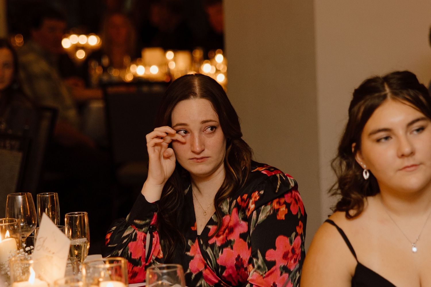 Two women seated at candlelit dinner table wearing elegant evening attire in warm ambient lighting.