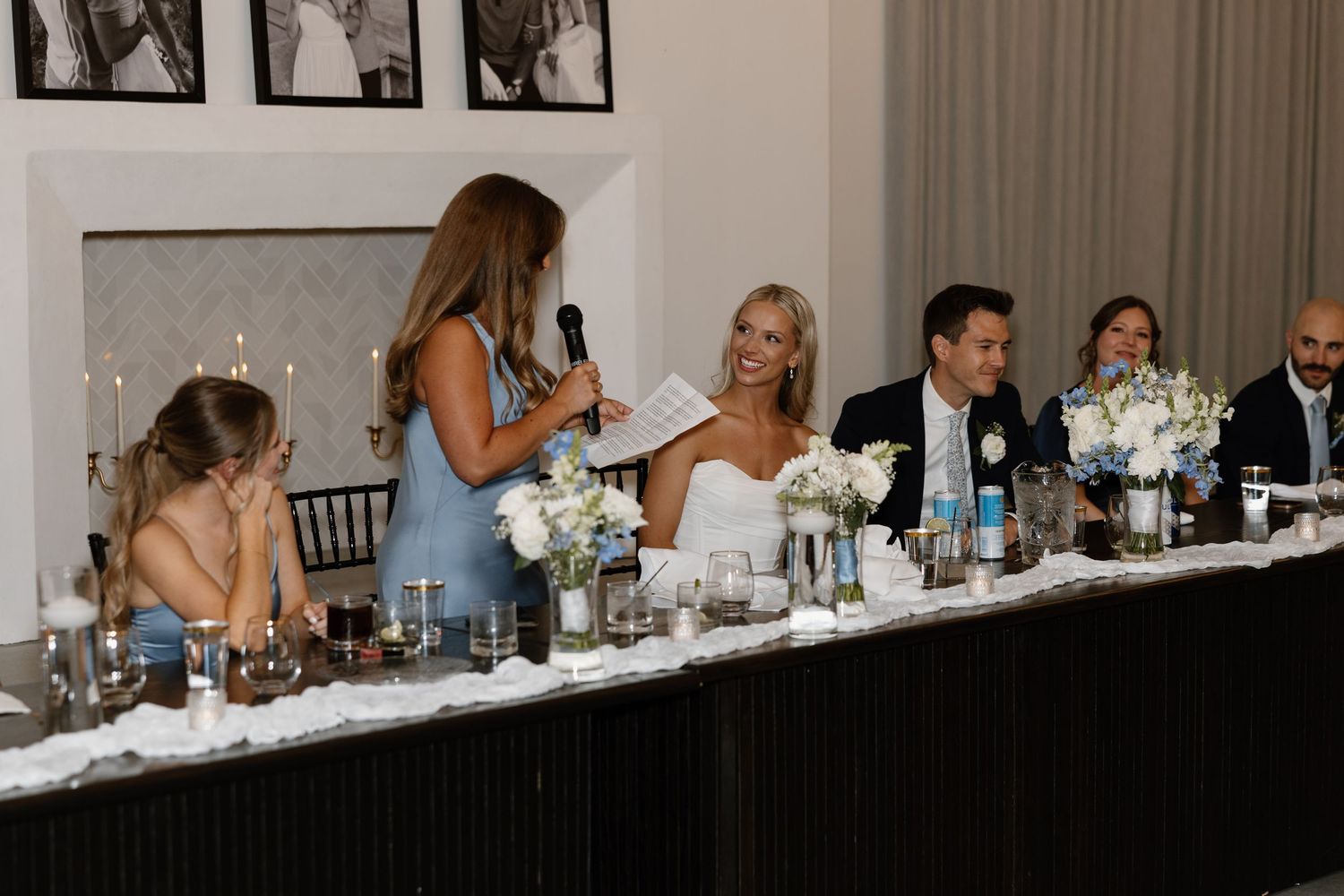 Wedding party sits at a long head table during reception dinner while guests socialize and celebrate.