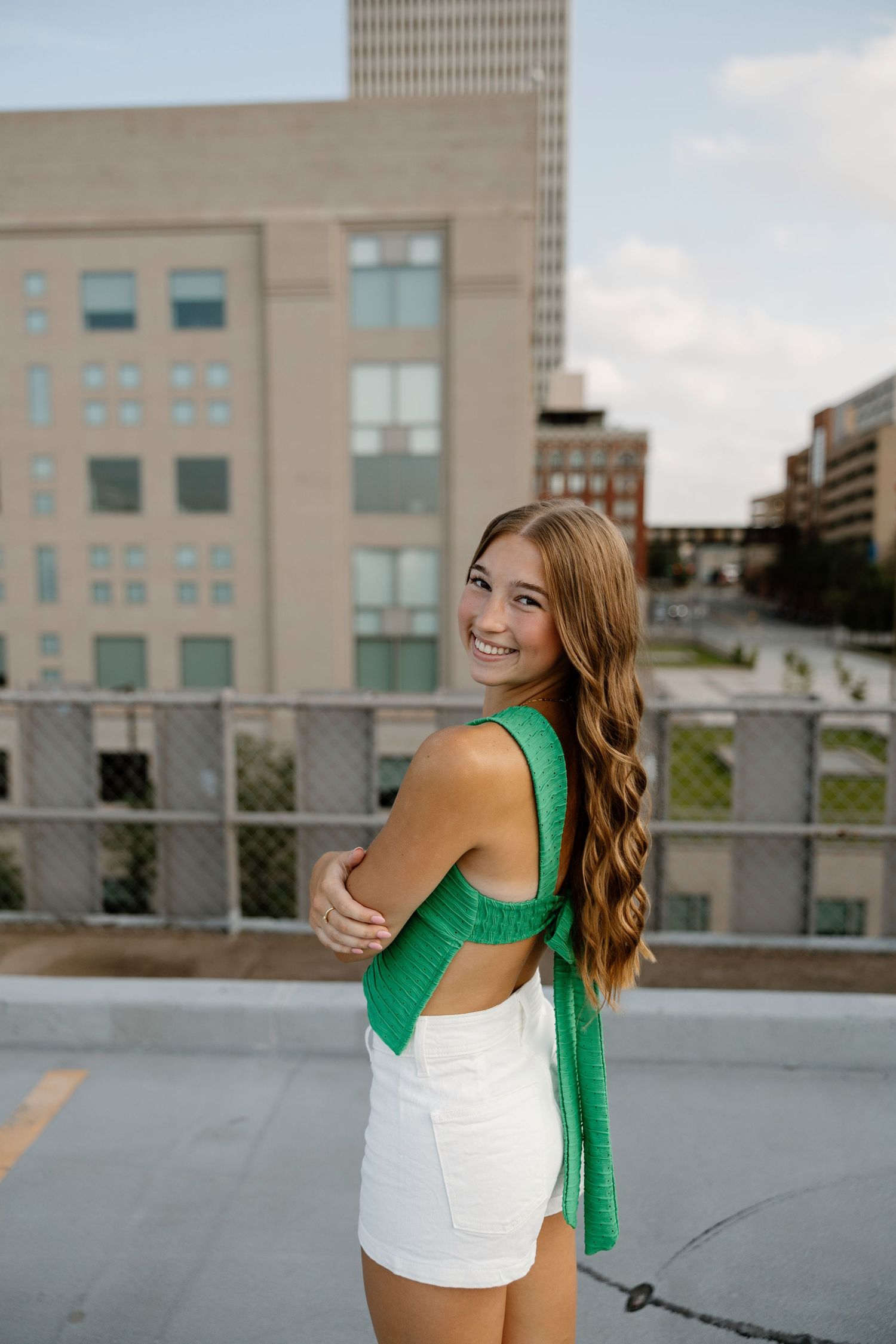 Woman in green cardigan and white top twirls joyfully on an urban rooftop with city buildings in background.